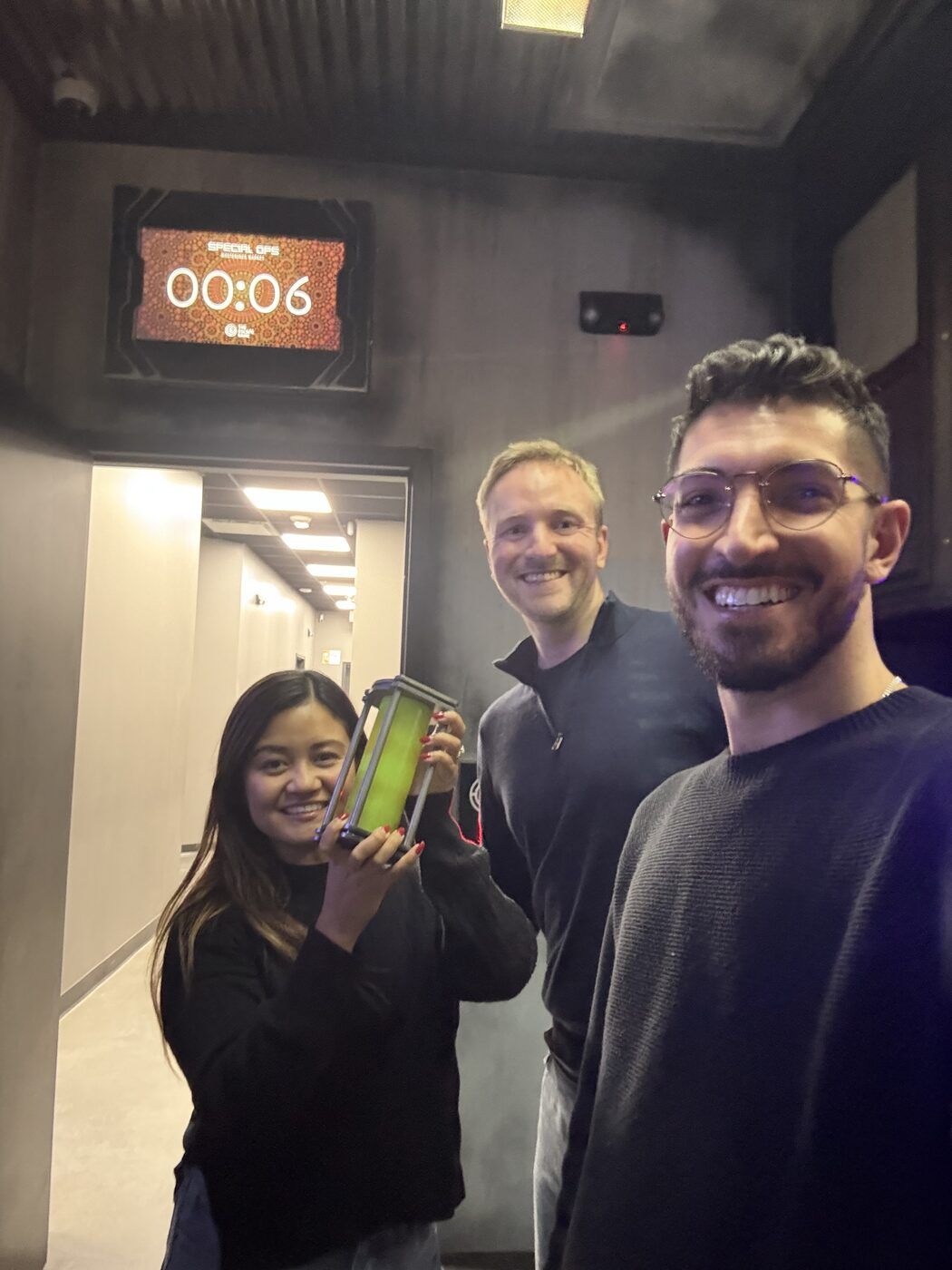 Three people smiling in an escape room corridor beneath a timer reading 00:06