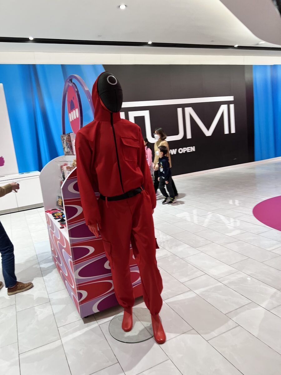 A figure in a red Squid Game guard costume standing beside a promotional booth