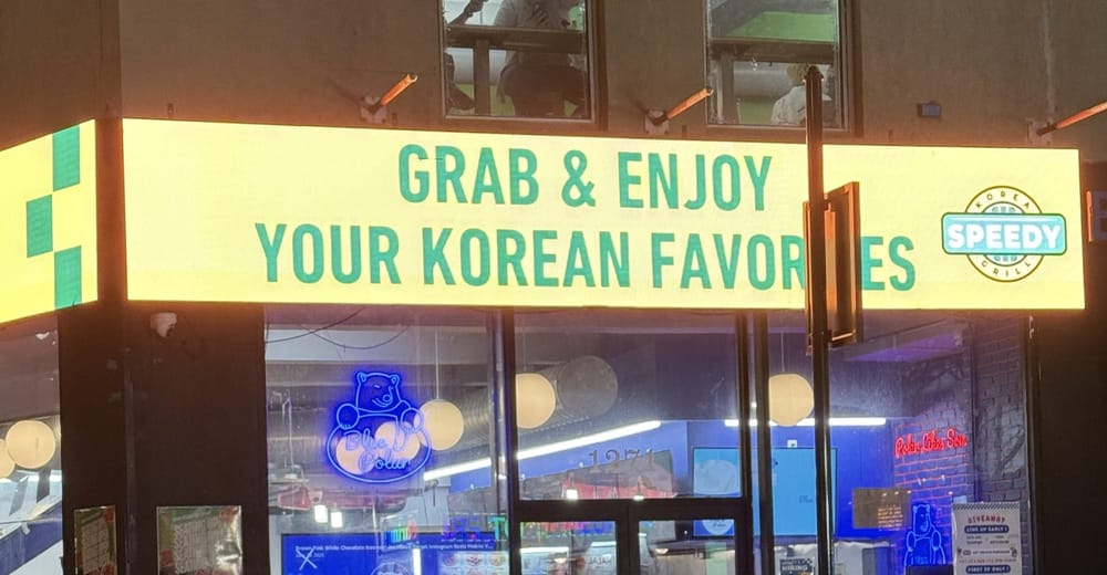 The glowing yellow Grab and Enjoy Your Korean Favorites sign above Speedy Korea Grill on Broadway in Koreatown at night