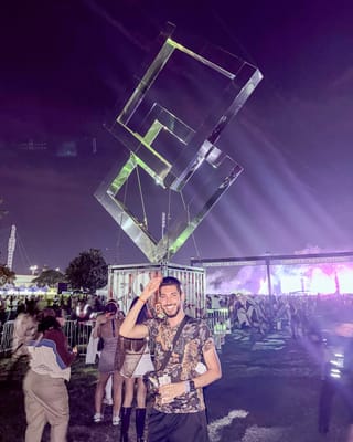 Rocky saluting and grinning in a tiger-print shirt under a giant hanging sculpture of clear interlocking squares, with the Experts Only Festival arch and the glowing main stage behind him at night