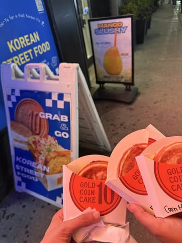 Three gold coin cakes in red Gold Coin Cake 10 sleeves held up on the sidewalk outside Speedy Korea Grill