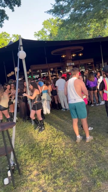 A black stretch tent on the grass with a wooden bar and a hanging chandelier ring, people crowding in