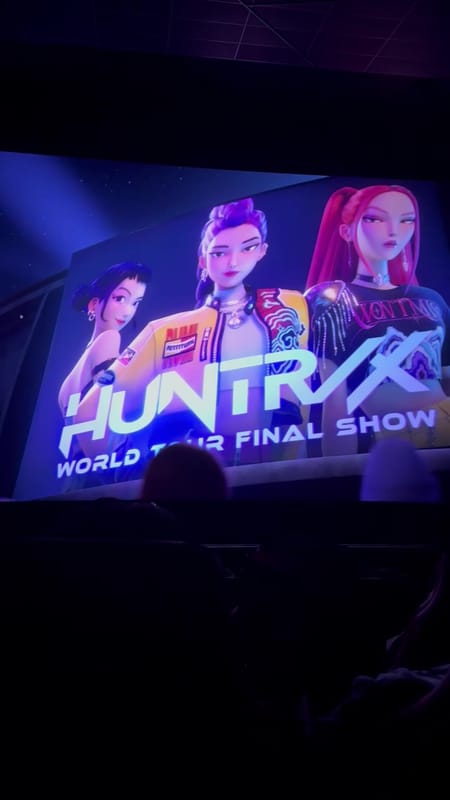 A huge HUNTR/X World Tour Final Show title card on the RPX screen, with dark rows of seats in the foreground
