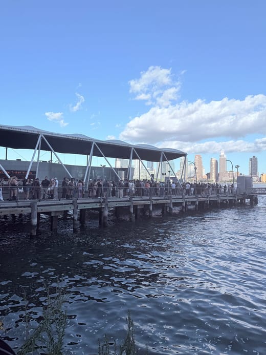 A long line of festival-goers along a wooden ferry pier under a white canopy, with the river in front and city towers under a blue sky