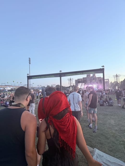 Tiffany in a red headscarf and Vinny walking across the lawn toward a black arch reading Experts Only Festival, with stage lights glowing at dusk