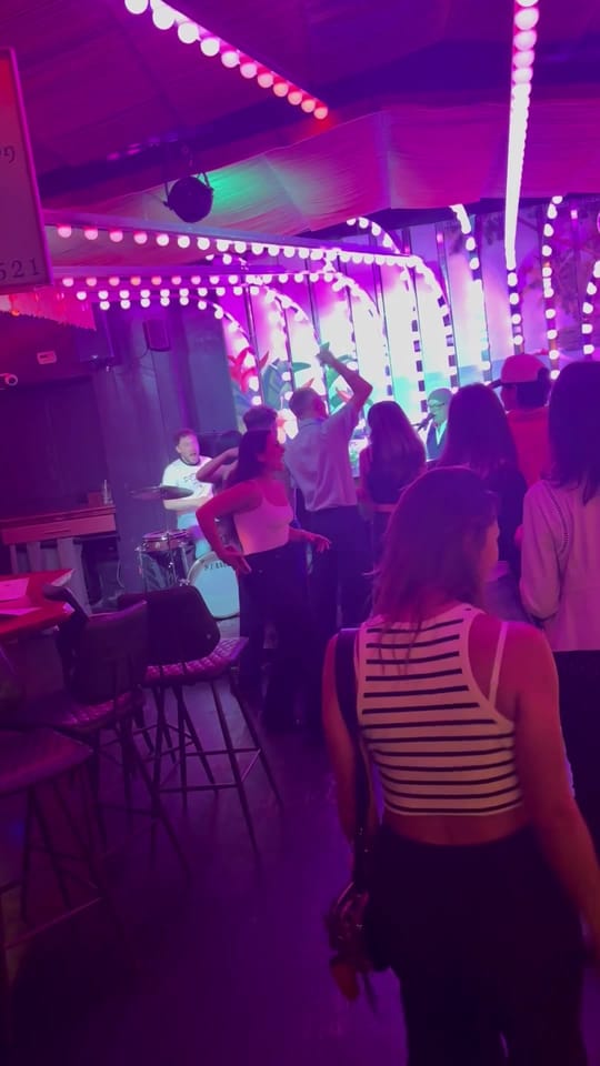 A crowd dancing in front of the stage at The Friki Tiki under pink and purple light strings