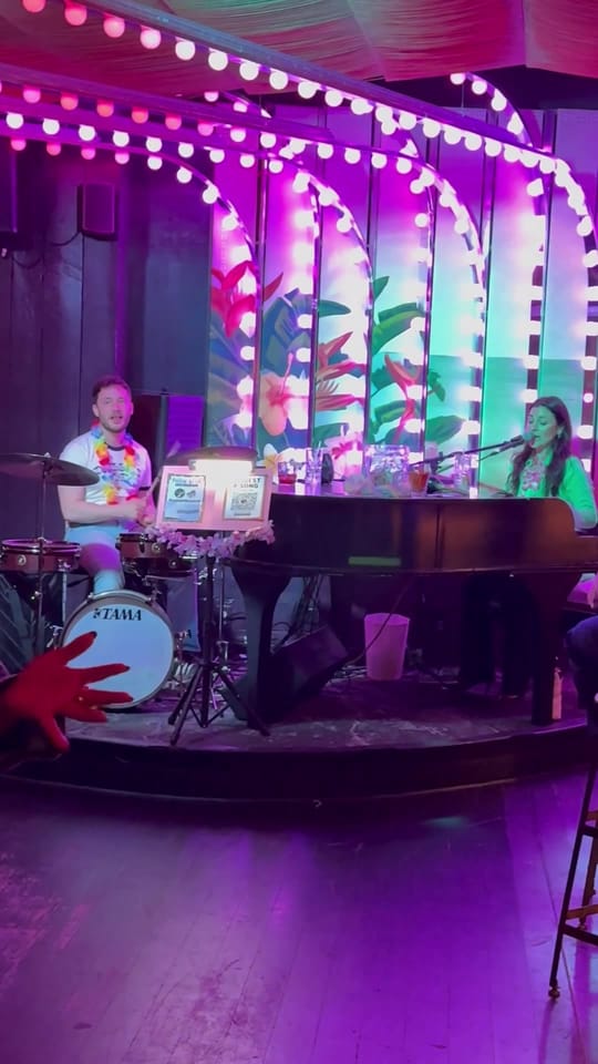Samantha Sayah at the piano and a drummer on the lit-up stage at The Friki Tiki