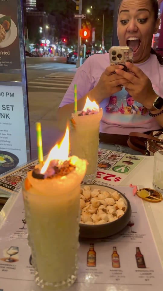 Carolyn filming with her phone and gasping as two flaming coconut cocktails arrive at the table