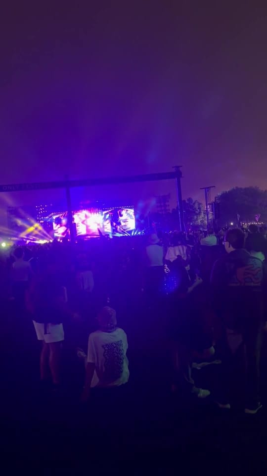 Walking from the green sculpture toward the main stage, glowing purple through the haze over a crowd on the grass