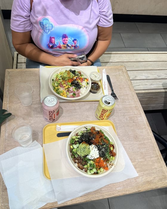 Carolyn at a red poke table in a lavender KPop Demon Hunters t-shirt, with two poke bowls on yellow trays in front of her