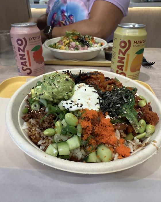 Rocky's red poke bowl up close: bulgogi beef, spicy pork, avocado, a poached egg, masago, seaweed, edamame and cucumber over rice, with cans of lychee and mango Sanzo behind it