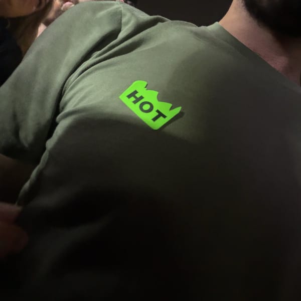 A neon green sticker that says HOT stuck to the shoulder of Rocky’s olive T-shirt in the crowd