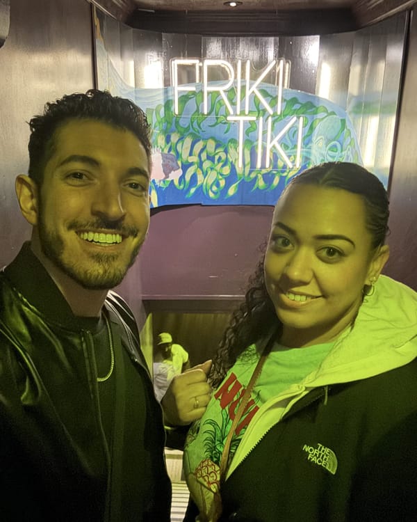 Rocky and Carolyn grinning under the Friki Tiki neon sign and wave mural at the entrance