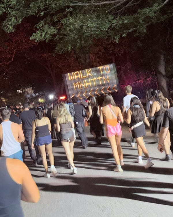 A crowd walking out of the festival at night past an orange LED road sign reading Walk to Mnhttn with arrows pointing left