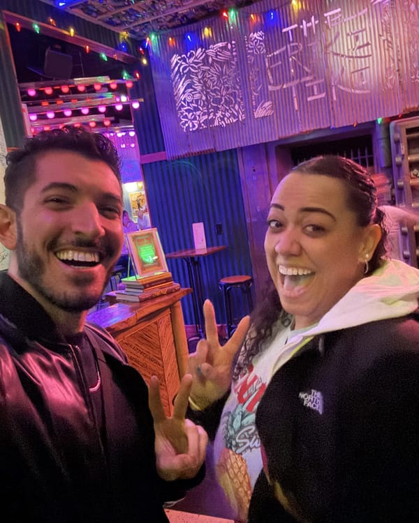 Rocky and Carolyn laughing inside The Friki Tiki, Carolyn making a peace sign, with the tiki bar sign lit up behind them