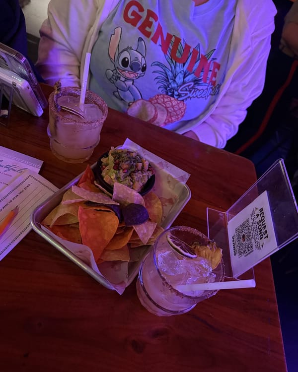 Two Aloha-Rita cocktails with spiced rims next to a tray of tortilla chips and Frikimole guacamole, Carolyn in a Stitch sweatshirt behind them