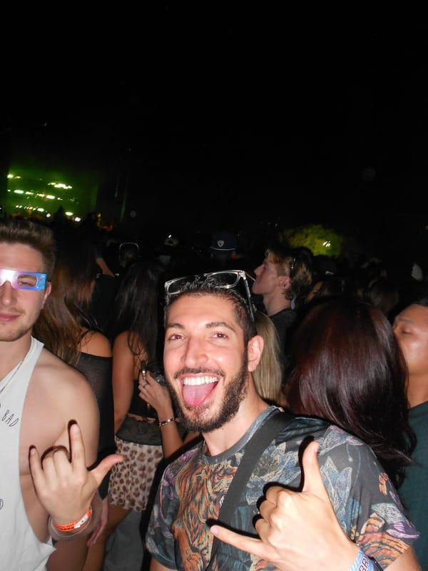 Zach in paper glasses with colored rims beside Rocky, who is sticking his tongue out and throwing a shaka, lit by a camera flash in the crowd