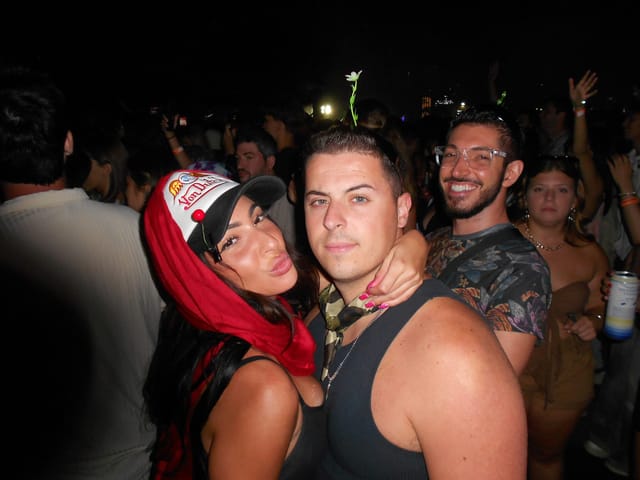 Tiffany in a trucker hat and red scarf, Vinny and Rocky posing together in the crowd at night, lit by a camera flash