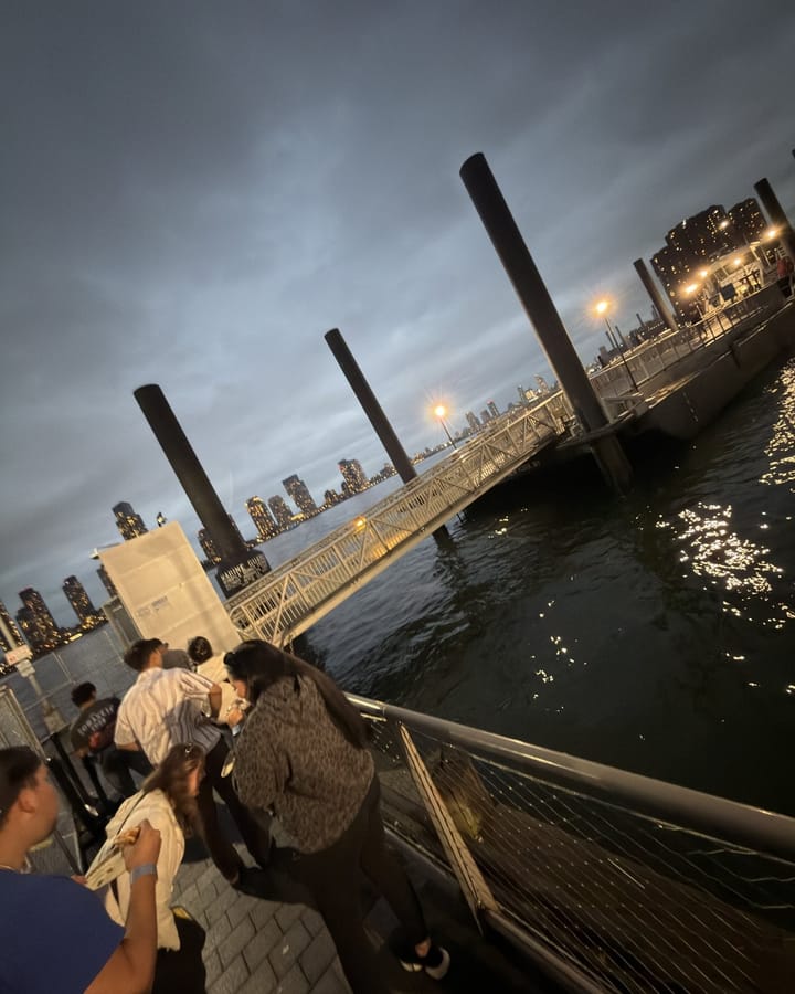 A handful of people on the East 35th Street ferry landing at dusk under a grey sky, the river and the lights of the waterfront behind them