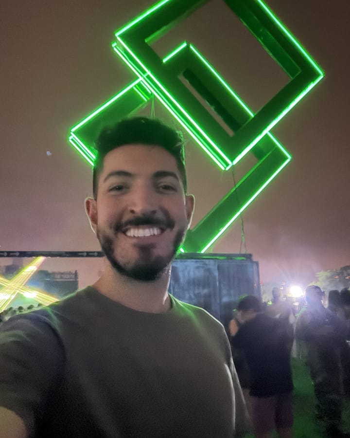 Rocky grinning in an olive T-shirt under a giant glowing green sculpture of interlocking squares at night, with lasers and the crowd behind him