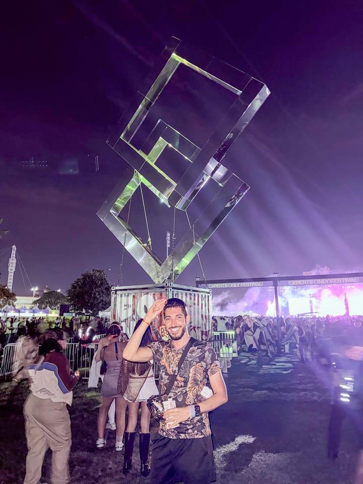 Rocky saluting and grinning in a tiger-print shirt under a giant hanging sculpture of clear interlocking squares, with the Experts Only Festival arch and the glowing main stage behind him at night