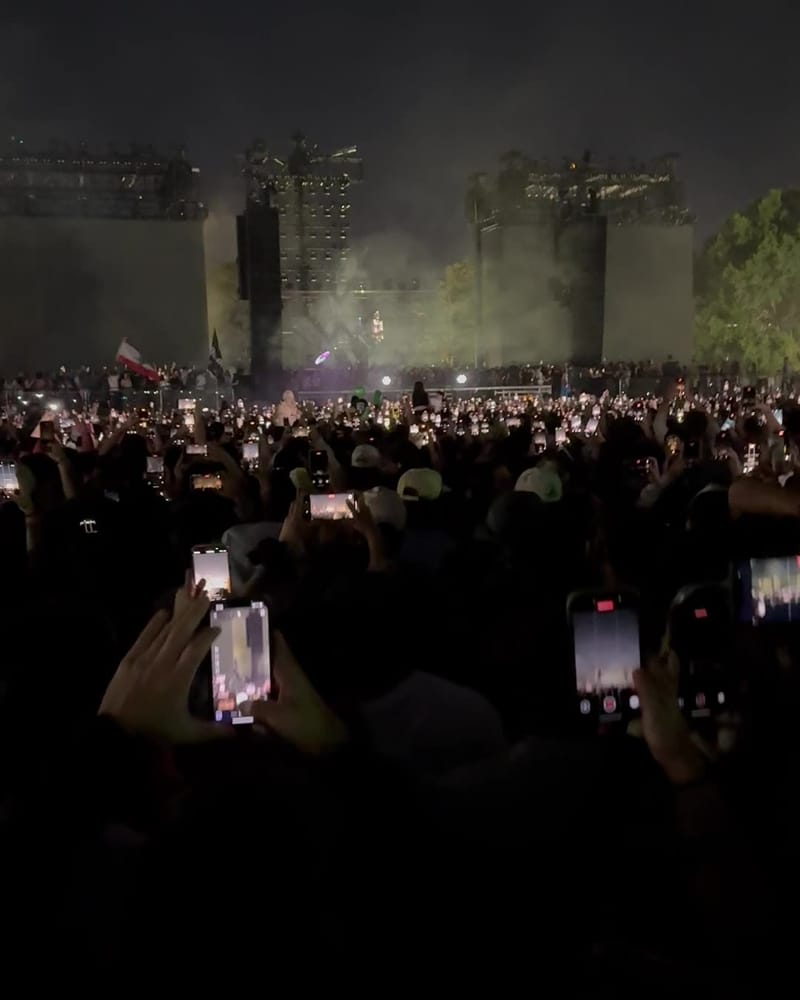 The main stage at night seen from inside the crowd, phone screens glowing all across the field under drifting smoke