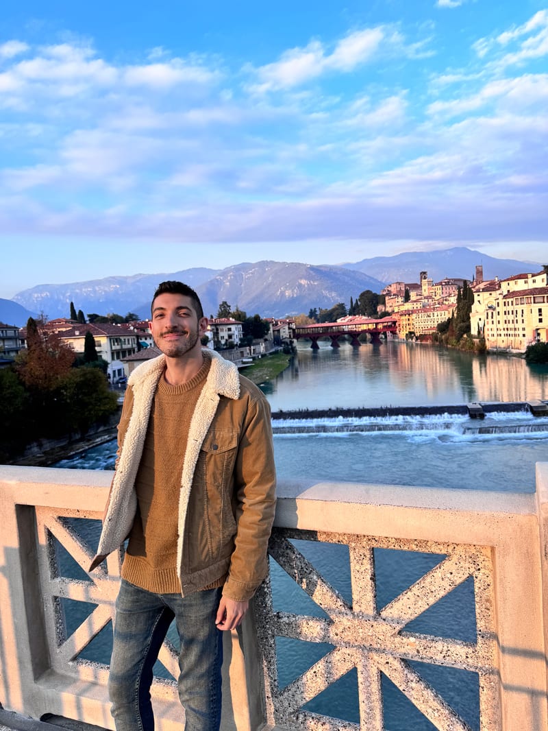 Rocky Trifari on a bridge over a river in northern Italy, a hilltop town and mountains behind him