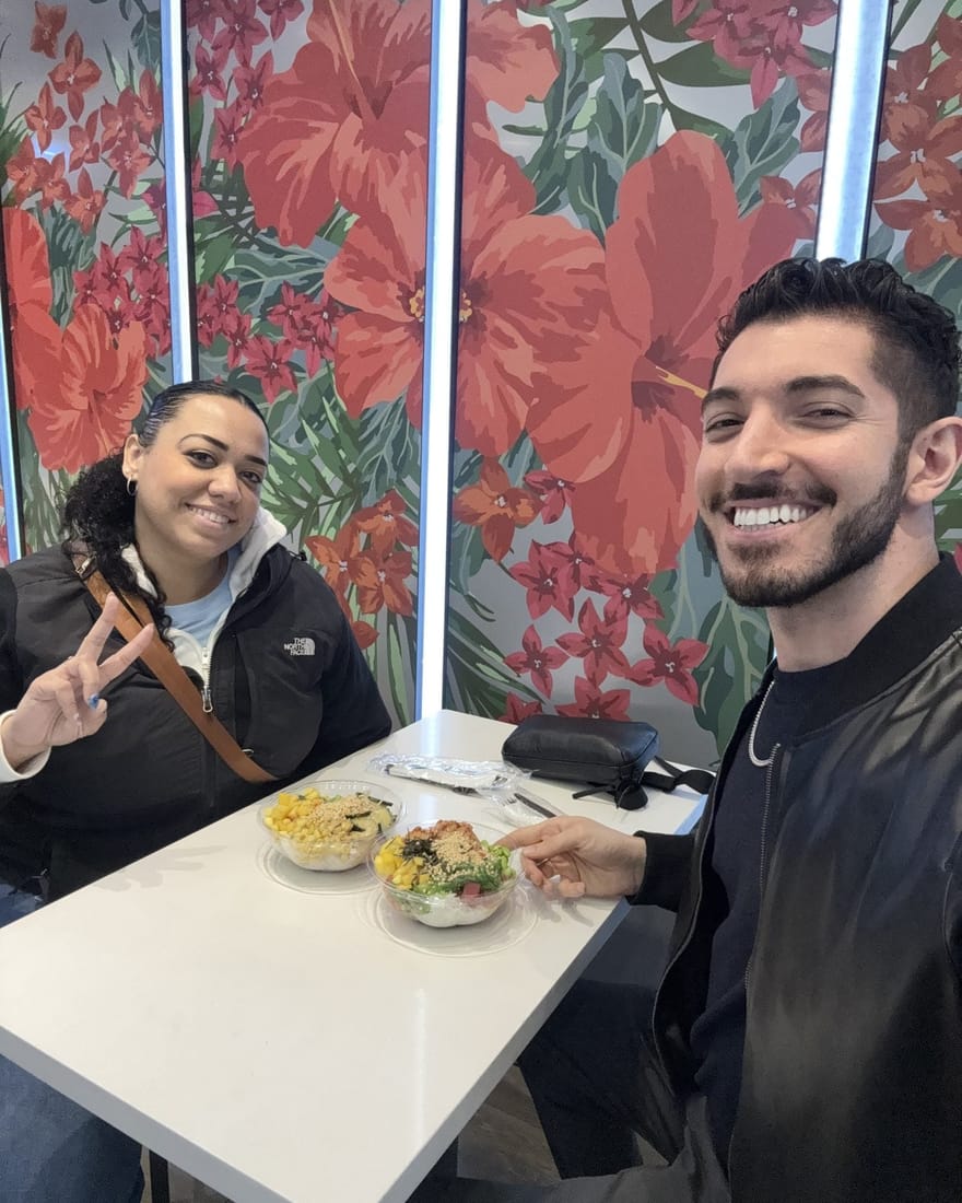 Rocky and his friend Carolyn smiling at a white table with two poke bowls, in front of a wall mural of giant red hibiscus flowers at Poke Bowl on West 39th Street