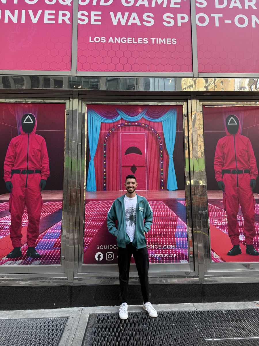 Rocky in a teal player tracksuit outside the Squid Game: The Experience entrance, flanked by two pink guards